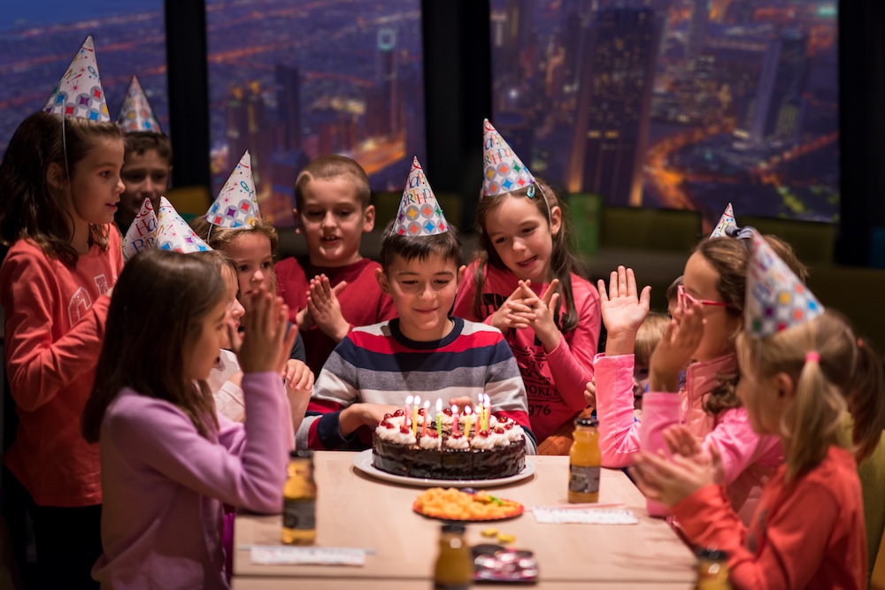 young happy boy and group of his friends having birthday party with a night city through the windows in the background